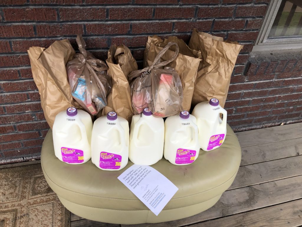 Six bags of groceries and five gallons of milk are stacked on a bench on a porch in front of a brick house.